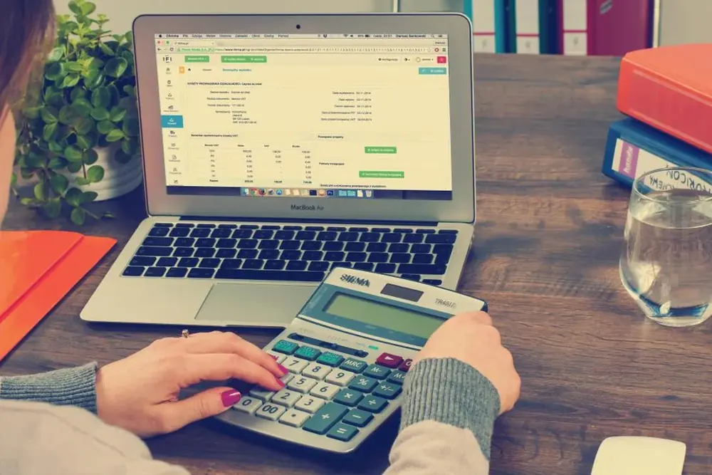 Hands using a calculator beside a laptop showing a ledger.